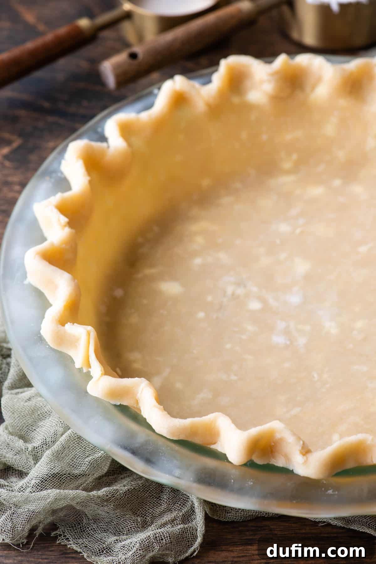 Gluten-free pie crust in a glass pie dish on a wooden surface, with a light green towel and measuring cups of ingredients in the background, showcasing a finished, unbaked crust.