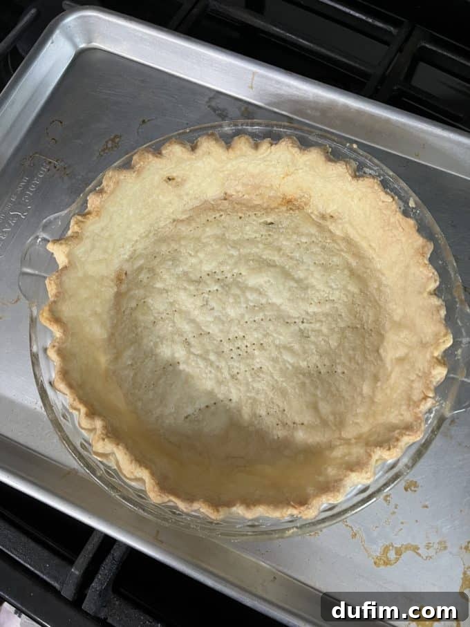 A gluten-free pie crust partially baked in a clear glass pie dish, sitting on a silver baking tray, showing signs of improvement.