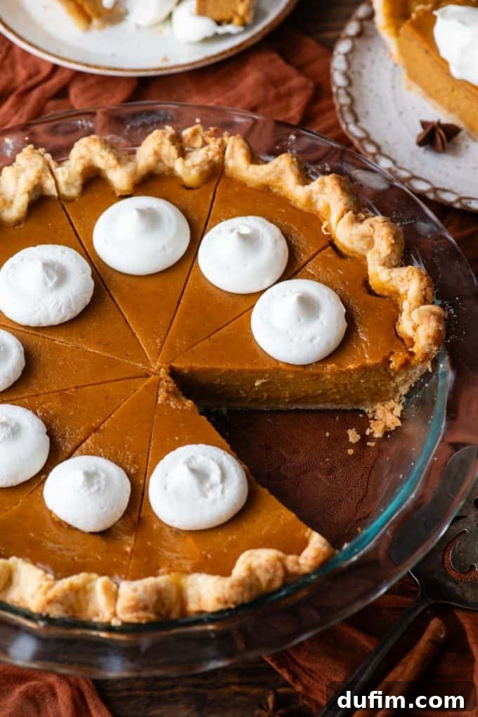 A freshly baked pumpkin pie in a glass pie dish, adorned with dollops of whipped cream, with one slice missing, set on a dark orange cloth on a wooden surface, with two plates holding pie slices beside it.