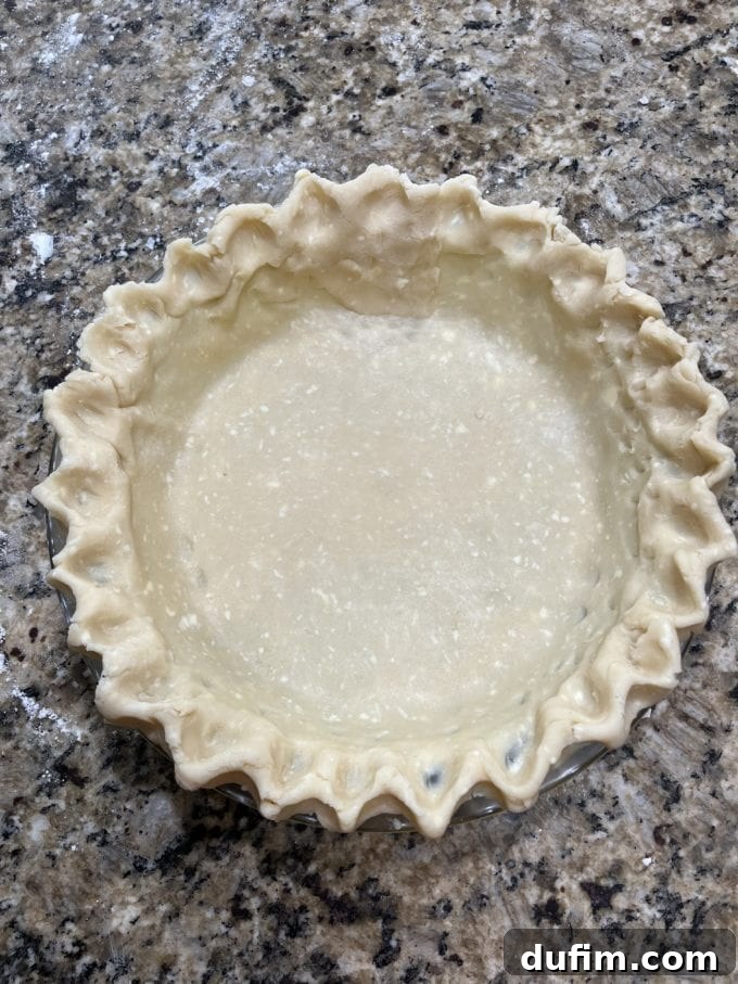 A gluten-free pie crust in a glass pie dish on a granite countertop, with edges being practiced for fluting.