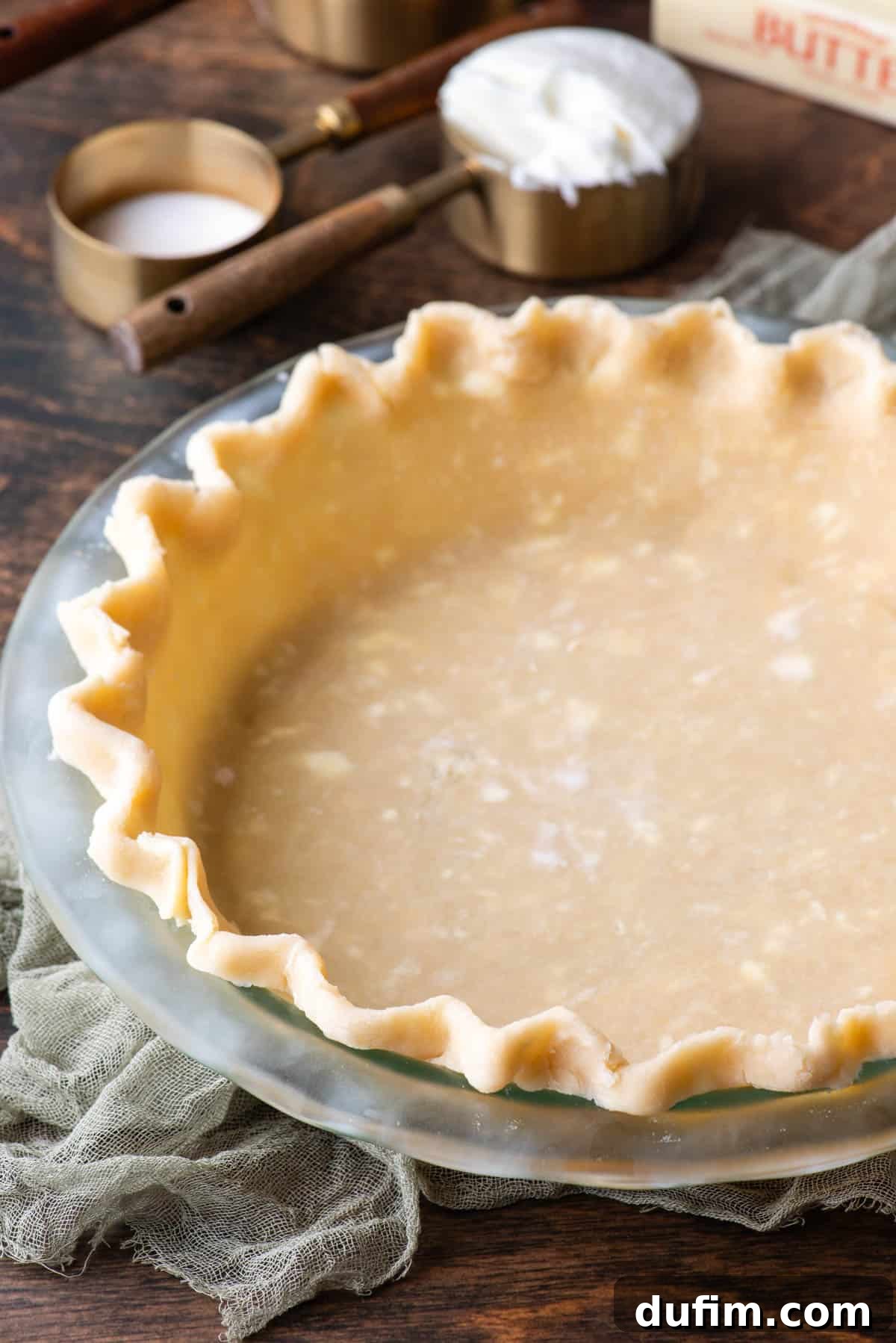 Gluten-free pie crust in a glass pie dish on a wooden surface, surrounded by measuring cups filled with the essential ingredients like flour, butter, and shortening, with a light green towel nearby.