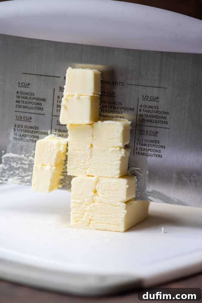 Cold butter being precisely cut into small cubes on a white cutting board using a pastry cutter, demonstrating the first step in preparing fats for the pie crust.