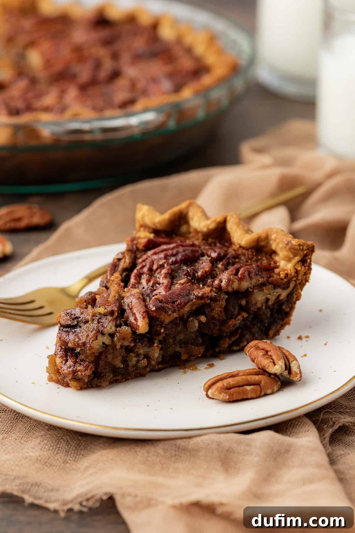 A perfectly baked slice of pecan pie, showcasing its rich, gooey filling and golden-brown crust, served on a white plate with a fork. Whole pecans are scattered around, resting on a tan towel and a rustic wood surface, with the full pie dish blurred in the background.