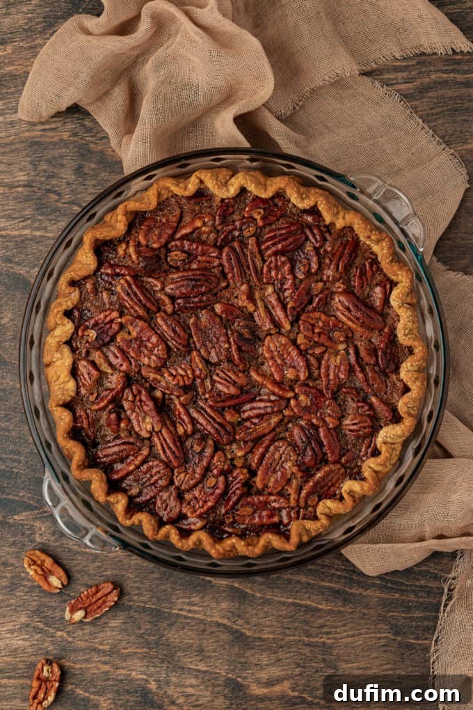 A glorious freshly baked pecan pie, golden and glistening, presented in a glass baking dish. It sits on a warm wood surface with a soft tan towel and whole pecans artfully scattered around, creating an appetizing and cozy display.