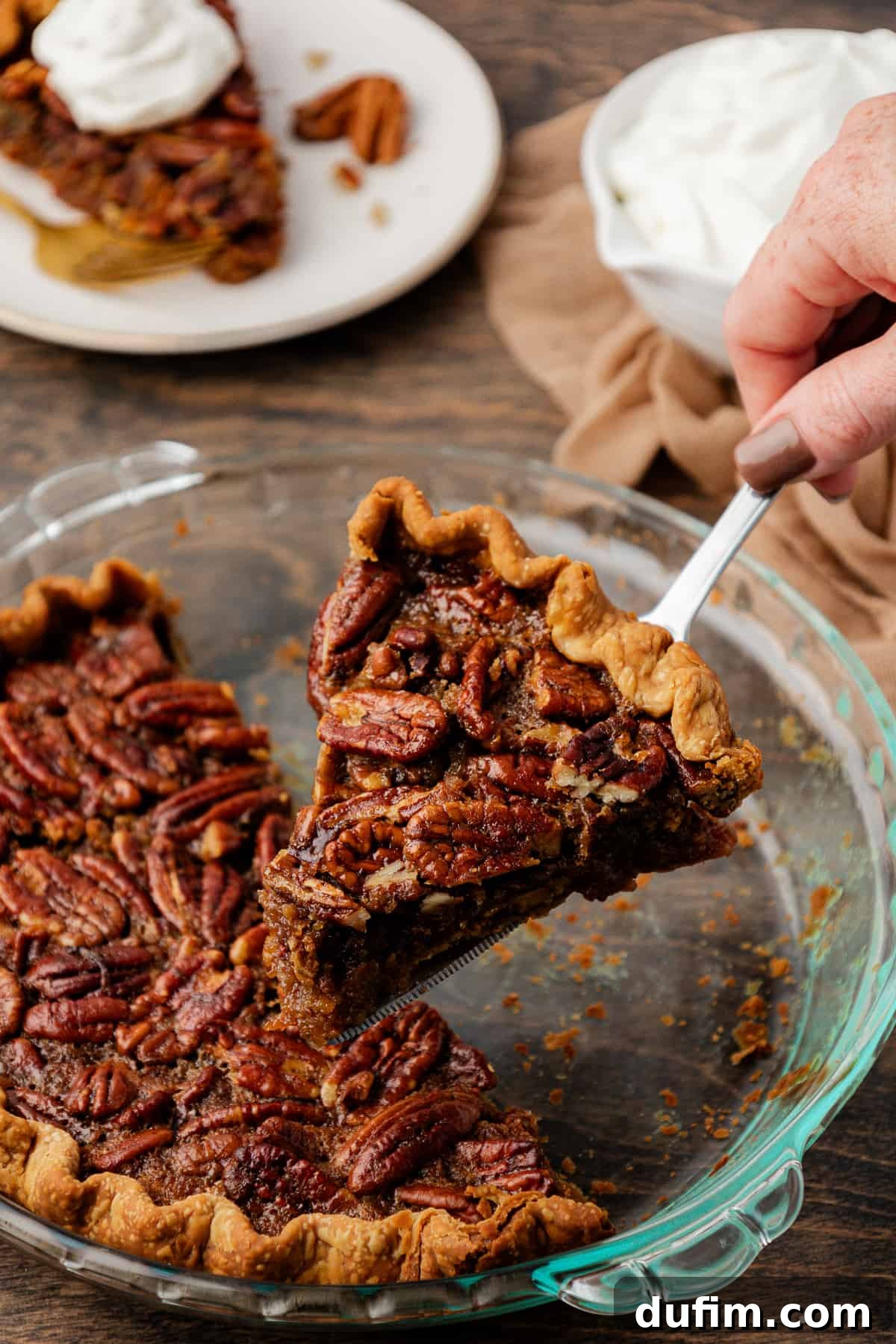 A tempting piece of pecan pie being gently lifted from a partially empty glass pie dish, which sits on a wood surface beside a soft tan towel. A nearby plate holds another slice, beautifully adorned with a dollop of whipped cream, and a small dish of whipped cream awaits eager diners. This arrangement suggests a delightful serving of freshly baked dessert.