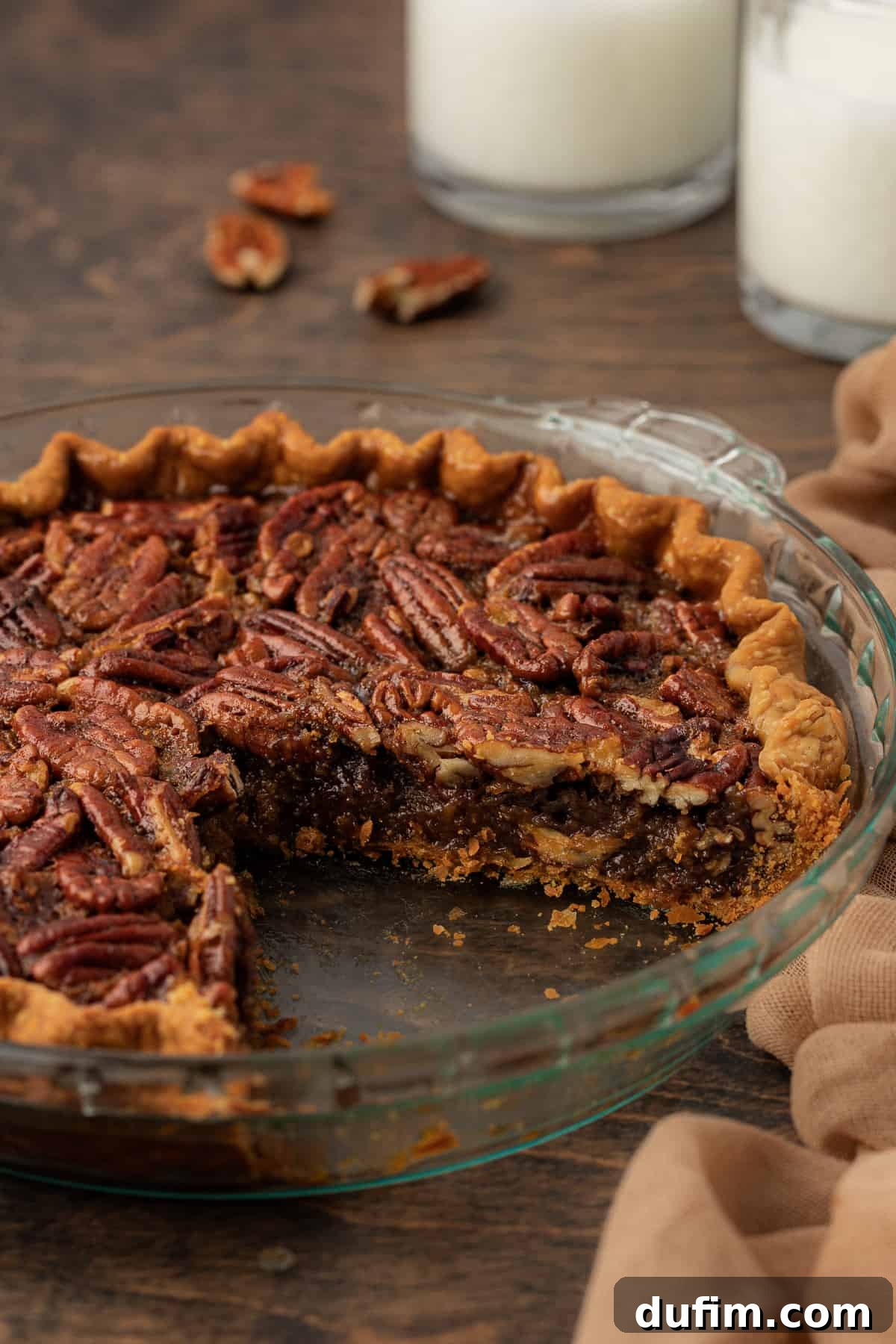 A mouth-watering pecan pie with a large, inviting slice already missing, showcased in a clear glass pie dish. It sits gracefully on a rich wood surface, surrounded by perfectly scattered whole pecans and a soft tan towel, with two glasses of milk subtly blurred in the background, creating a cozy and enticing dessert scene.