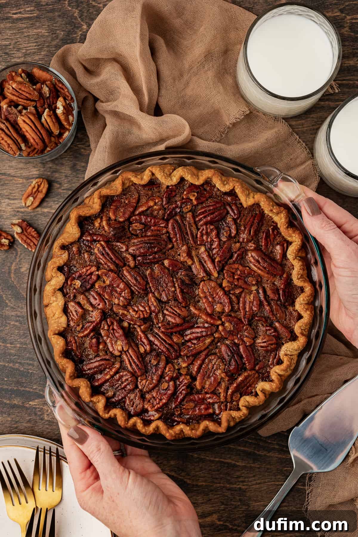 A freshly baked whole pecan pie being carefully placed onto a warm wood surface with two hands. It rests on a soft tan towel, surrounded by charming rustic elements: two glasses of milk, a small dish overflowing with pecans, more loose pecans scattered artfully, a neat stack of plates and forks, and a pie serving spatula, all creating a cozy, inviting scene.