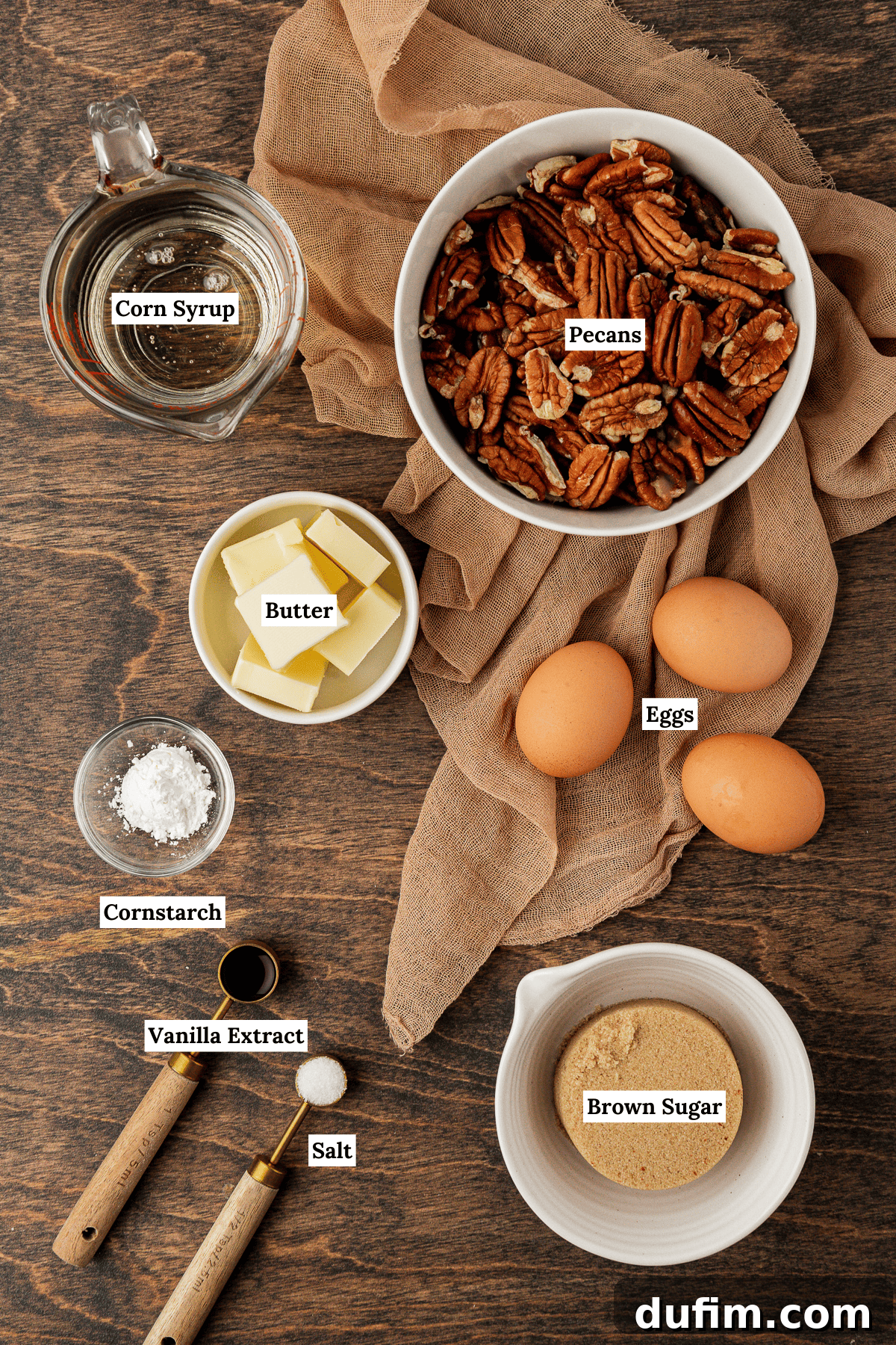 A beautifully arranged spread of essential pecan pie ingredients on a rustic wood surface, featuring a tan towel as a backdrop. The ingredients include clear corn syrup, whole pecans, a stick of butter, fresh eggs, a small dish of cornstarch, a bottle of vanilla extract, a pinch of salt, and a mound of brown sugar, all perfectly prepped for baking.