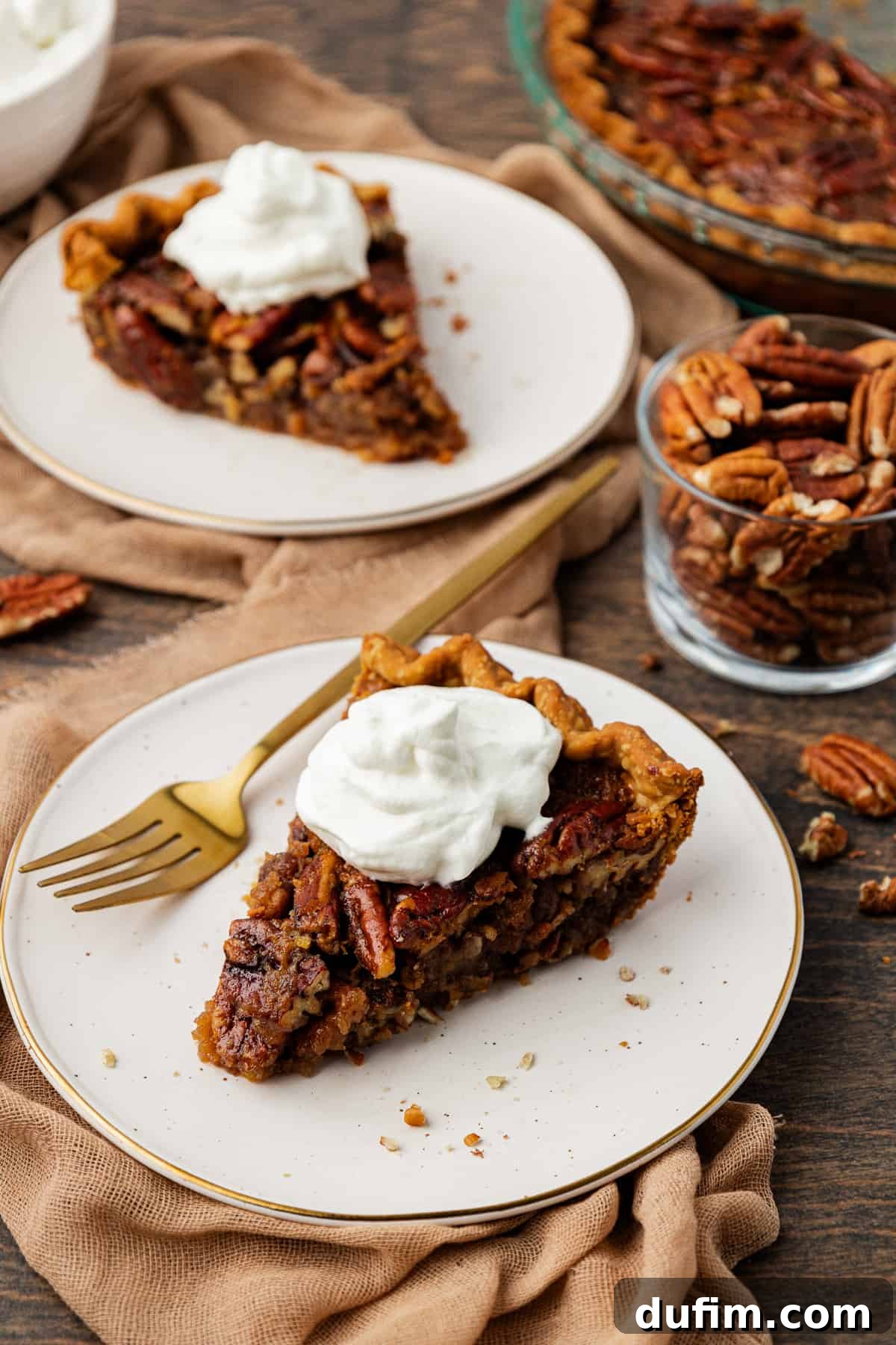 Two pristine white plates each hold a perfect slice of pecan pie, with a fork resting on the front plate, inviting you to indulge. They sit gracefully on a tan towel atop a warm wood surface, accompanied by a small glass dish brimming with whole pecans and more pecans artfully scattered around. In the softly blurred background, a glass pie dish containing more delicious pie completes the enticing scene.