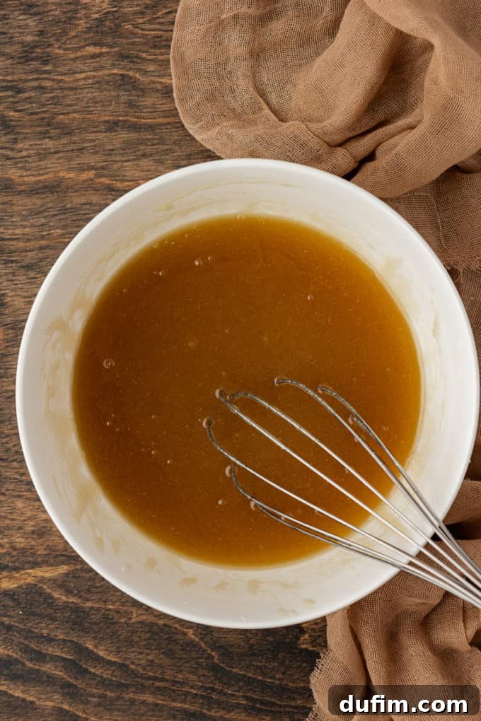 Wet ingredients for pecan pie being thoroughly whisked together in a pristine white bowl, set on a warm wood surface with a soft tan towel neatly folded beside it.