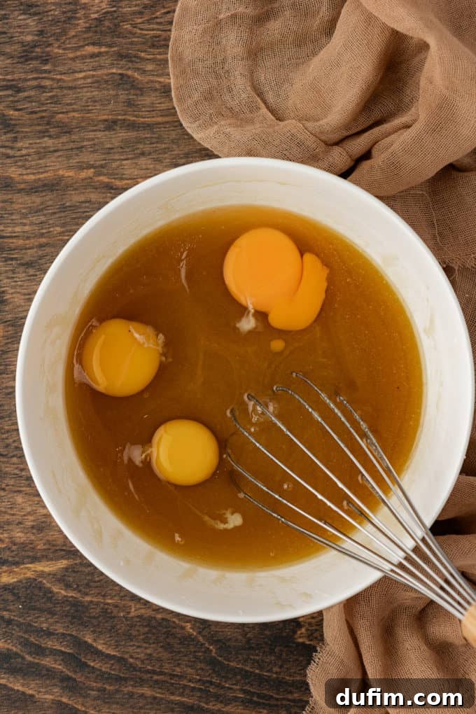 Wet ingredients for pecan pie being whisked together in a white bowl on a wood surface, with a tan towel nearby, showing a few unmixed egg yolks suspended in the liquid before full incorporation.