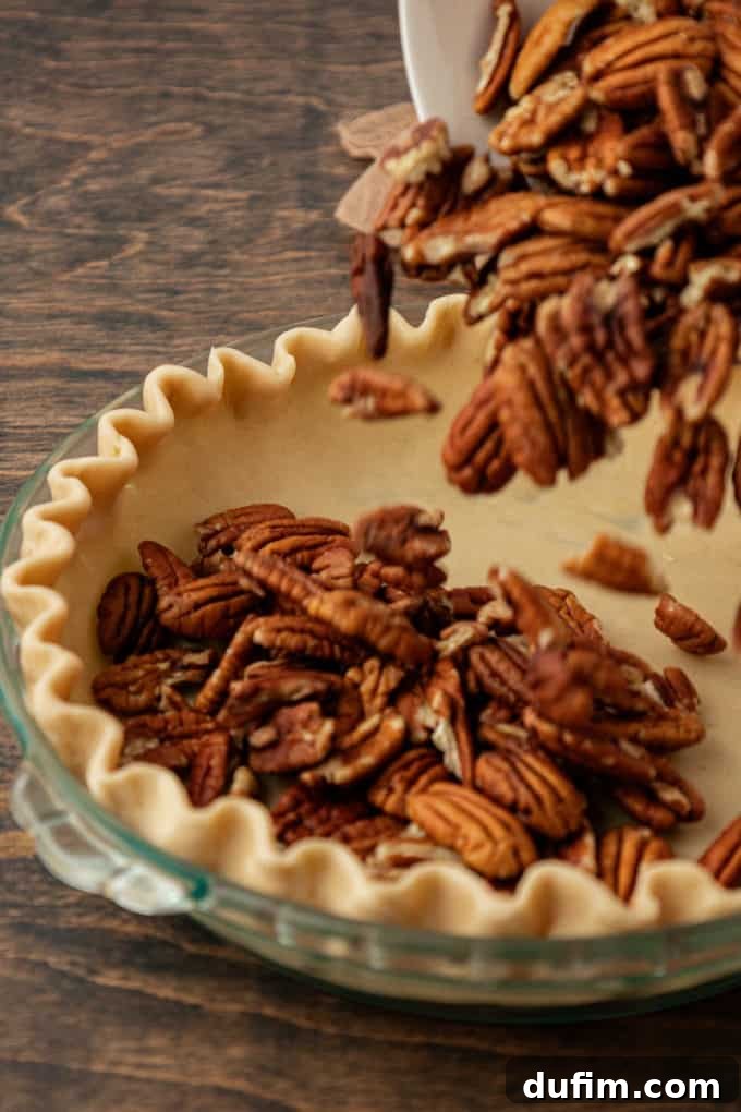 Whole pecans are being generously added to a clear glass pie dish, which already contains an unbaked pie crust, resting on a sturdy wood surface, ready for the filling.