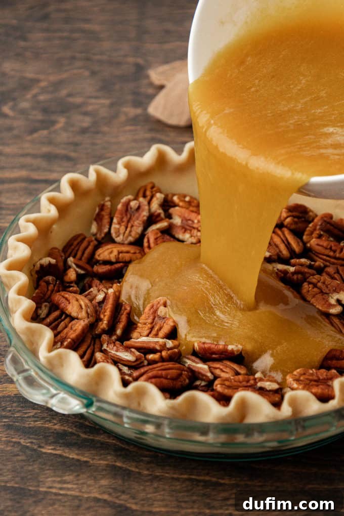 A clear glass pie dish filled with a generous layer of pecans sits on a wood surface, as the rich, amber pecan pie filling mixture is steadily poured over the nuts, preparing for baking.