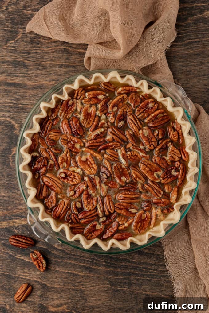 An unbaked pecan pie, perfectly assembled and ready for the oven, resting in a glass baking dish on a wood surface, framed by a tan towel and scattered whole pecans, creating a rustic and inviting culinary scene.
