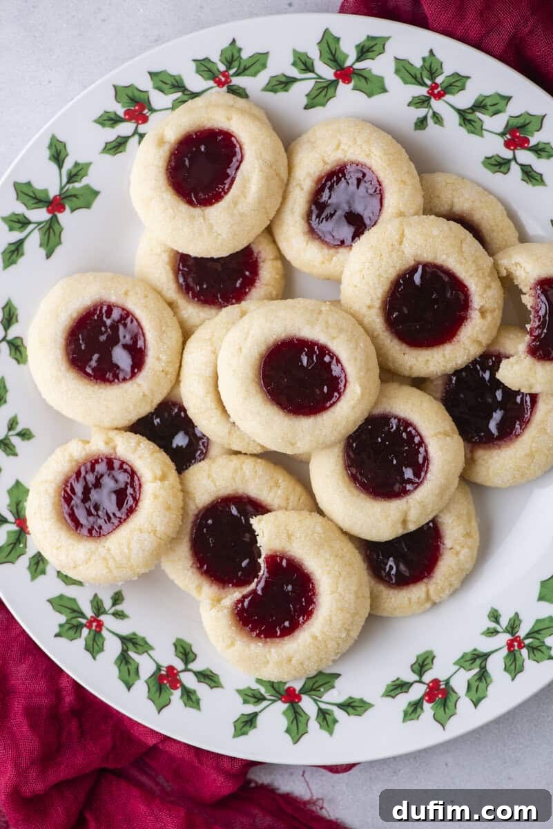 THUMBPRINT COOKIES: A TIMELESS DELIGHT 2 A festive plate decorated with holly holds several classic thumbprint cookies filled with red raspberry jam, arranged in a pile on a light surface with a red cloth partially visible. These perfect thumbprint cookies are ideal for holiday tables.