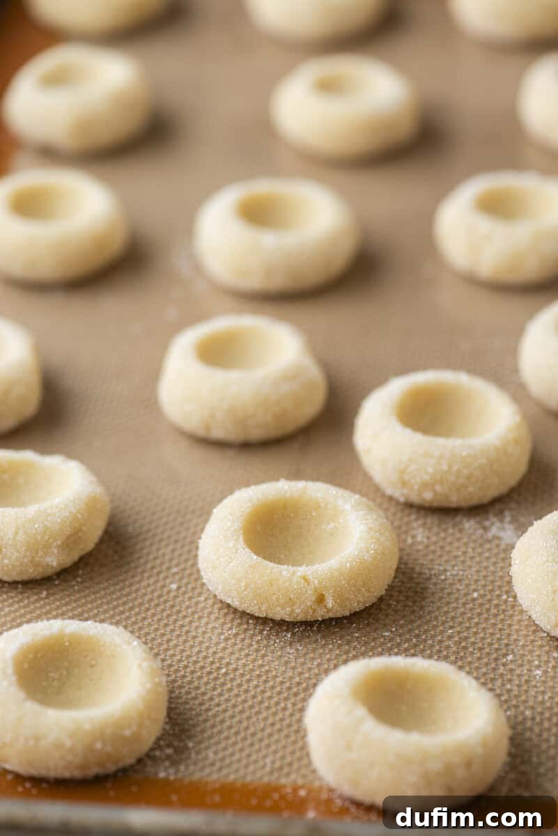 THUMBPRINT COOKIES: A TIMELESS DELIGHT 11 Rows of unbaked thumbprint cookies, each with a distinct indentation, are neatly arranged on a baking sheet lined with a silicone mat, coated lightly with sugar and spaced evenly apart.
