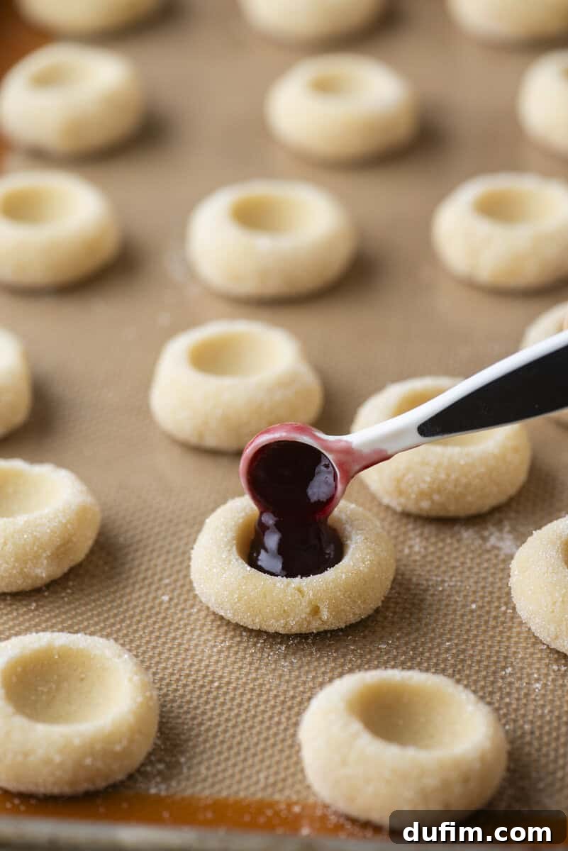 THUMBPRINT COOKIES: A TIMELESS DELIGHT 12 A hand uses a small spoon to meticulously fill the indented centers of delicious thumbprint cookies with vibrant red jam. Several unfilled cookies with perfect indents await their filling on a baking sheet lined with a silicone mat.