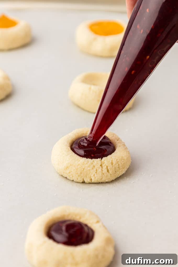 THUMBPRINT COOKIES: A TIMELESS DELIGHT 14 Thumbprint cookies on a baking sheet lined with parchment paper are being filled with bright red raspberry jam, ready for baking.
