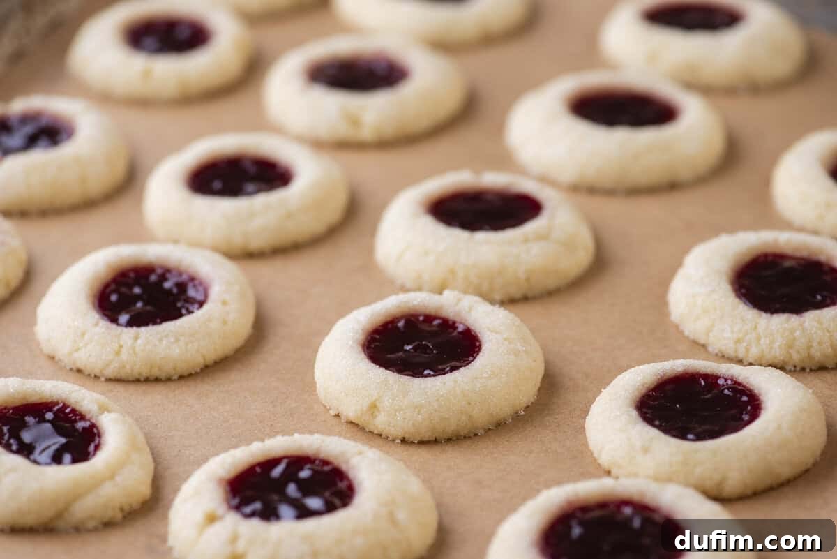 THUMBPRINT COOKIES: A TIMELESS DELIGHT 15 Rows of perfectly baked, round thumbprint cookies with bright red jam centers are arranged on a sheet of brown parchment paper. The crumbly, sugar-coated cookies are evenly spaced, showcasing their classic holiday charm and inviting texture.
