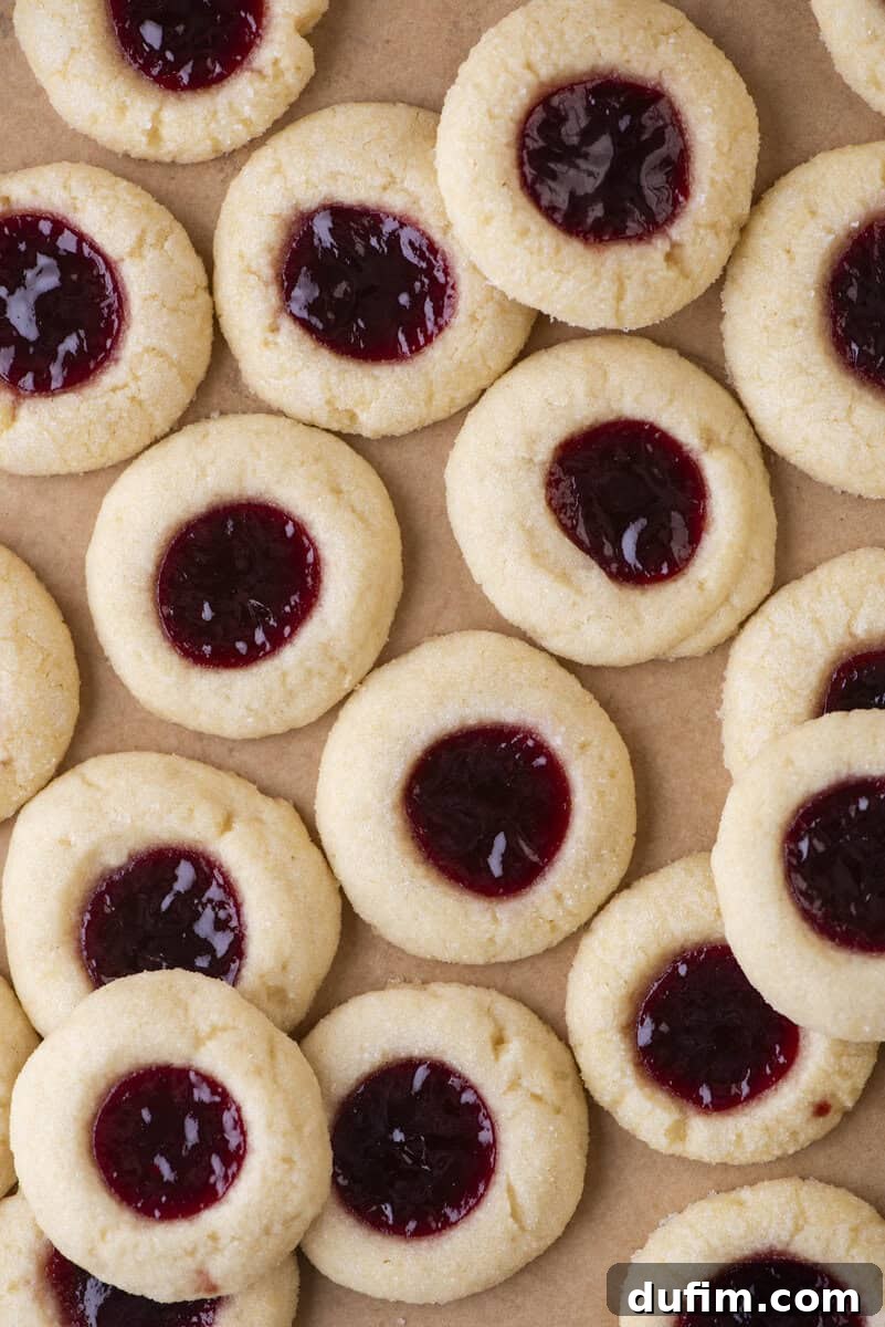 THUMBPRINT COOKIES: A TIMELESS DELIGHT 17 A close-up of several perfectly baked thumbprint cookies with a rich red jam filling in the center, arranged on brown parchment paper. The cookies display a soft, crumbly texture, indicative of a classic shortbread.