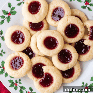 A plate decorated with holly displays a festive pile of classic jam thumbprint cookies. One cookie has a bite taken out, revealing its soft, tender texture and sweet raspberry jam filling.