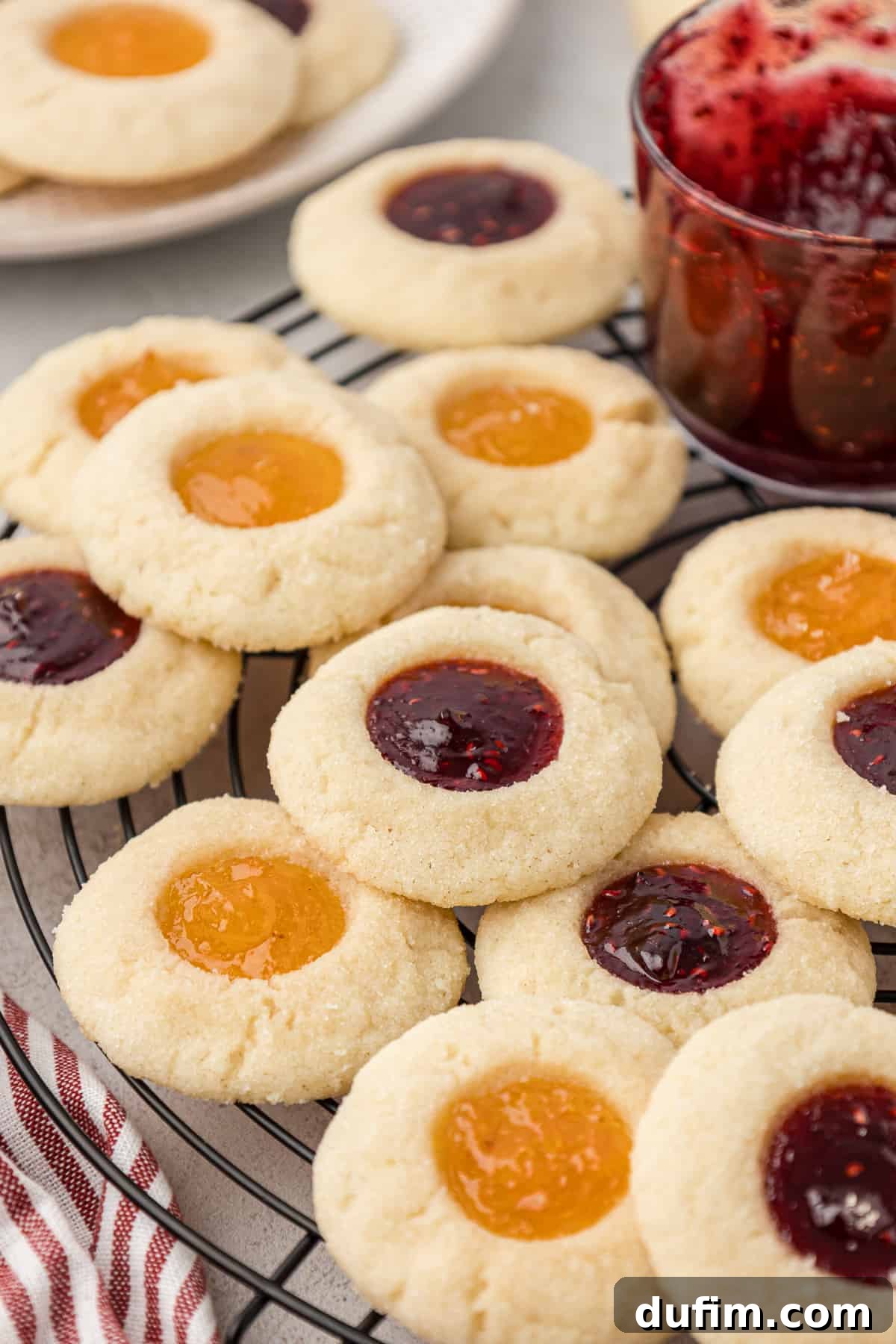 THUMBPRINT COOKIES: A TIMELESS DELIGHT 3 An assortment of apricot and raspberry jam thumbprint cookies are scattered on a wire cooling rack beside a jar of vibrant raspberry jam, highlighting their homemade charm and sweet filling.