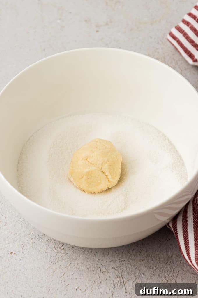 THUMBPRINT COOKIES: A TIMELESS DELIGHT 9 A perfectly formed ball of thumbprint cookie dough rests in a white bowl filled with granulated sugar on a countertop, ready for rolling.