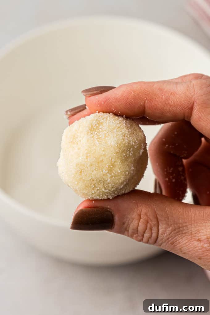 THUMBPRINT COOKIES: A TIMELESS DELIGHT 10 A hand holds a freshly sugar-coated ball of thumbprint cookie dough above a bowl of granulated sugar, demonstrating the uniform coating achieved.