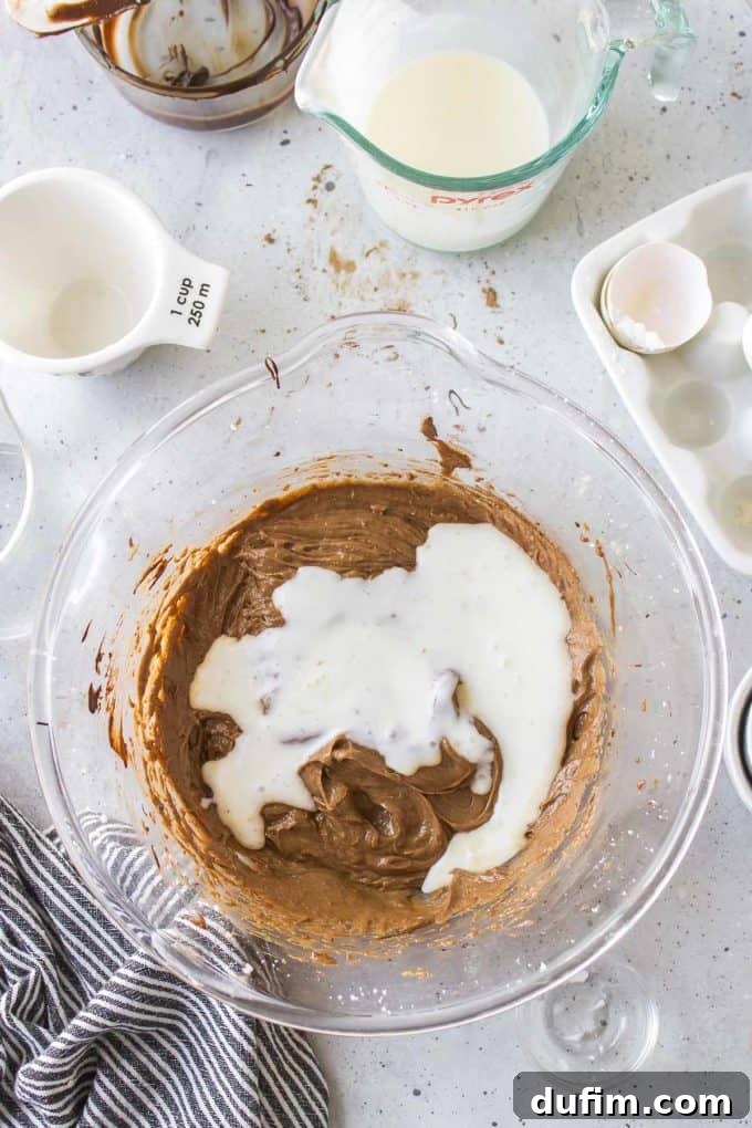 Ingredients for German chocolate cake in a glass bowl, accompanied by a black and white striped towel, a white measuring cup, an empty glass dish with chocolate remnants, a glass measuring cup of milk, and an empty eggshell. This setup shows the mixed wet ingredients before adding buttermilk.