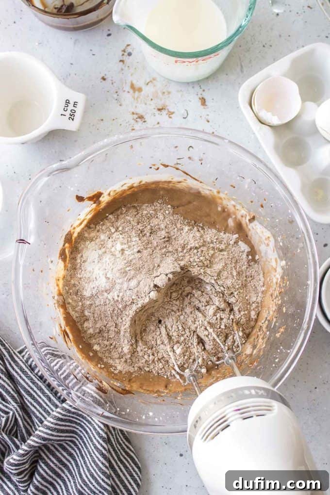 German chocolate cake ingredients being mixed in a glass bowl with an electric mixer. The setup includes a black and white striped towel, a white measuring cup, an empty glass dish with chocolate residue, a glass measuring cup of milk, and an empty eggshell, capturing the blending process.