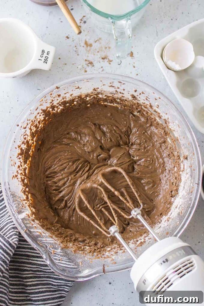 German chocolate cake ingredients actively mixing in a glass bowl with an electric mixer, surrounded by a black and white striped towel, a white measuring cup, a glass measuring cup of milk, and an empty eggshell, illustrating the final stages of batter preparation.