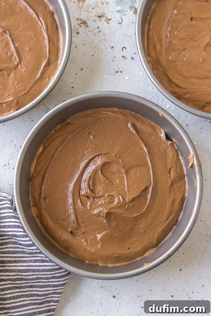 Three cake pans on a countertop, evenly filled with German chocolate cake batter, beside a black and white striped towel, ready for baking.