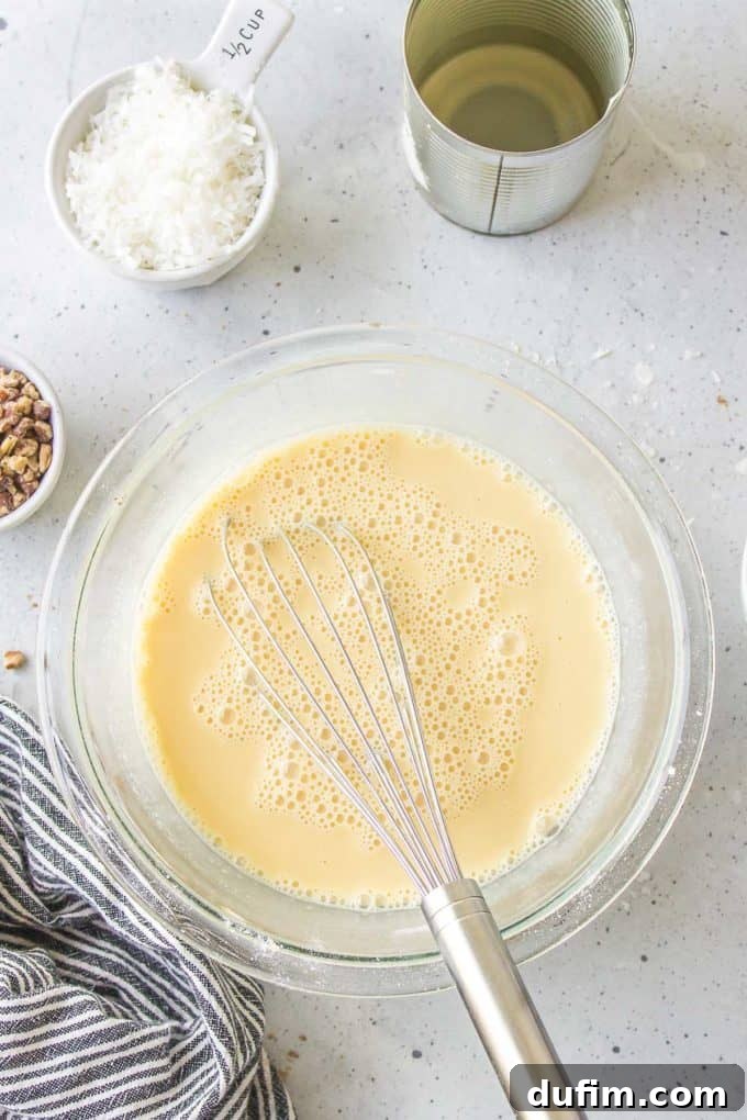 Wet ingredients for German chocolate cake frosting in a glass bowl with a whisk, positioned beside a black and white striped towel, a bowl of pecans, a measuring cup of shredded coconut, and an empty can with traces of evaporated milk, illustrating the initial mixing stage.