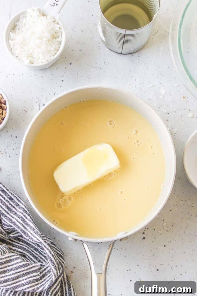 A piece of butter and the flour mixture for German chocolate cake frosting in a white pot, surrounded by a black and white striped towel, a small dish of pecans, a measuring cup of shredded coconuts, and an empty can of evaporated milk, preparing for the cooking process.