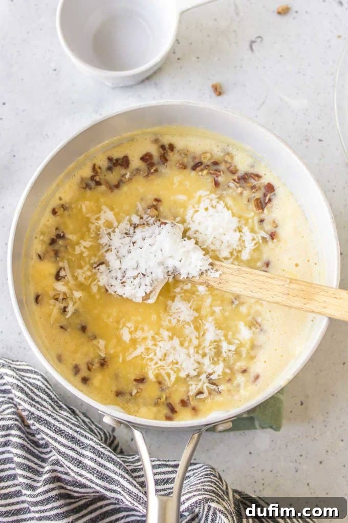 Ingredients for German chocolate cake coconut pecan frosting cooking in a white pot with a wooden spoon resting inside, accompanied by a black and white striped kitchen towel and an empty white bowl, showing the mixture thickening on the stove.