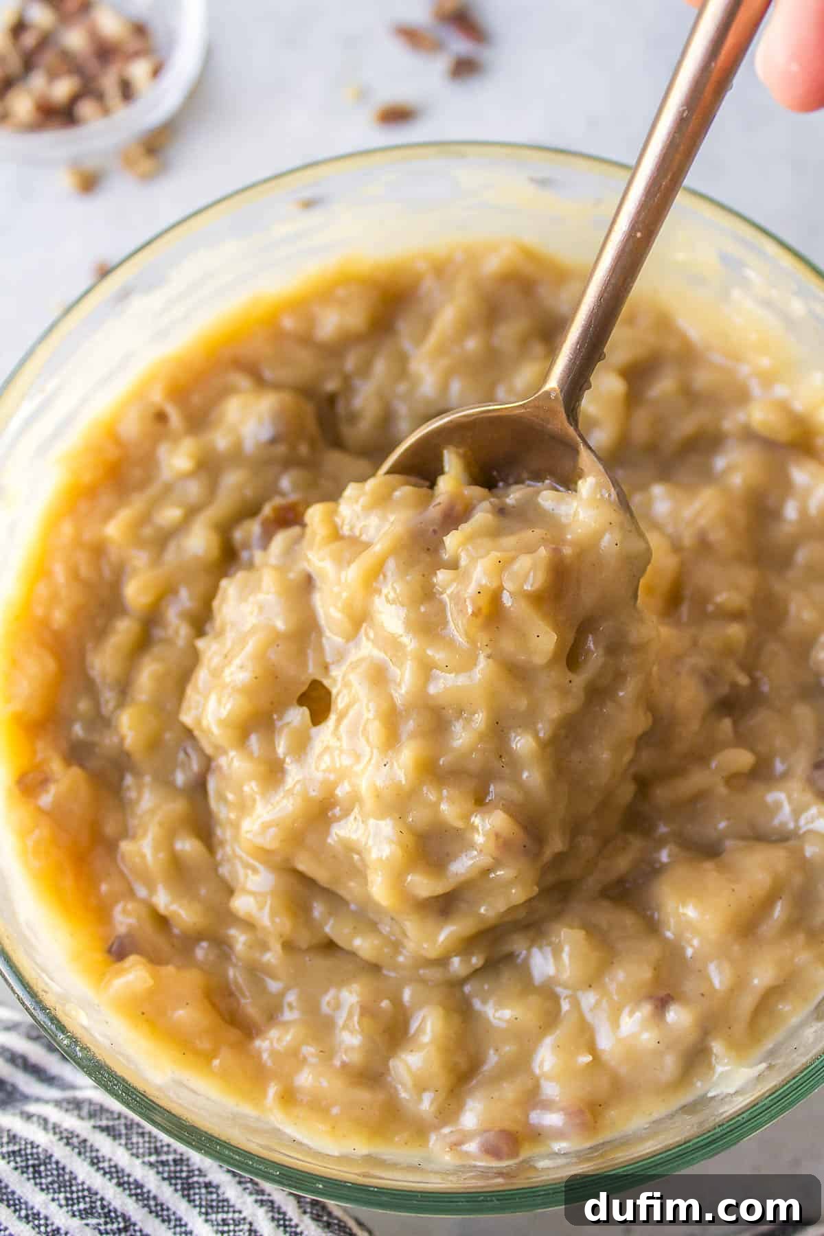 German chocolate cake coconut pecan frosting in a glass bowl with a spoon, ready for use, with a small dish of pecans blurred in the background, highlighting the rich, thick texture of the frosting.