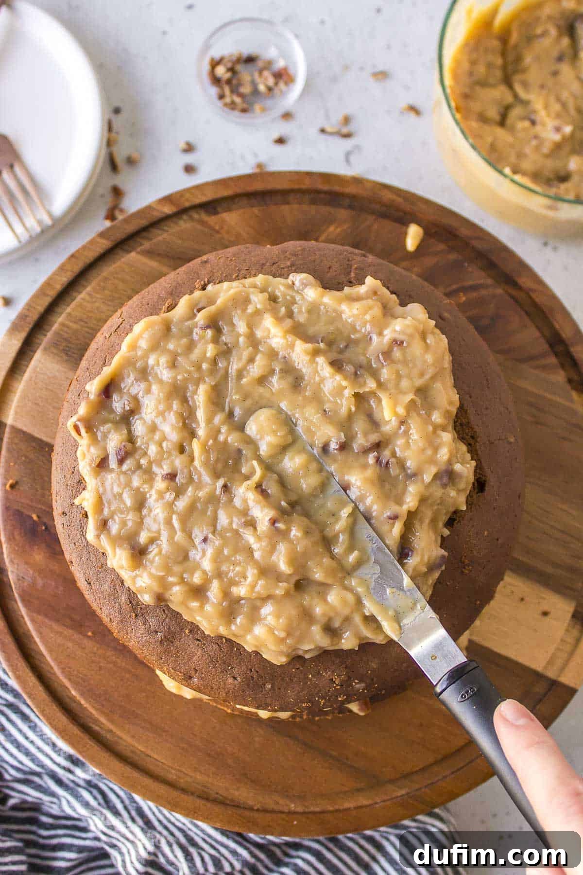 Coconut pecan frosting being carefully spread onto a layer of German chocolate cake, which rests on a wooden cake serving platter, showcasing the assembly process.