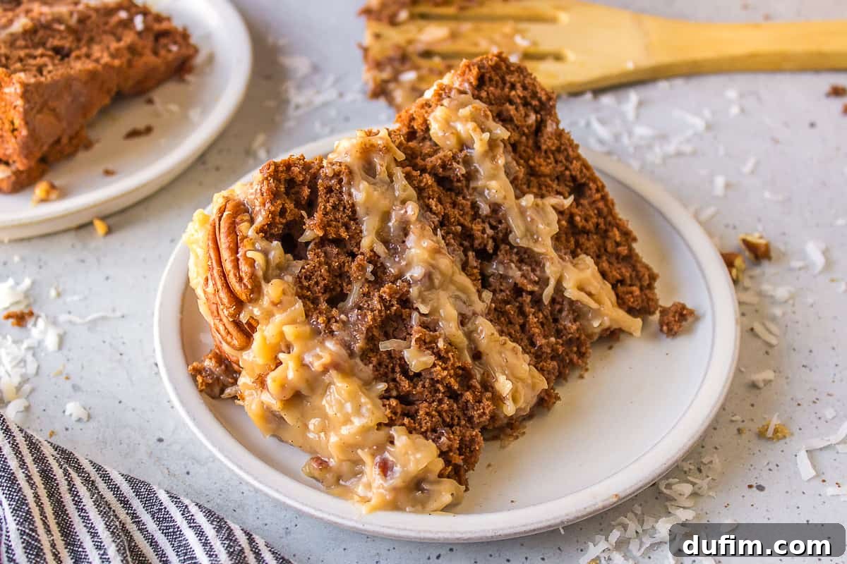 A delectable slice of German chocolate cake, artfully placed on its side on a white plate. It's surrounded by a charming black and white striped kitchen towel and sprinkled with shredded coconut. A wooden spatula with cake remnants and another slice in the background complete this inviting dessert display.