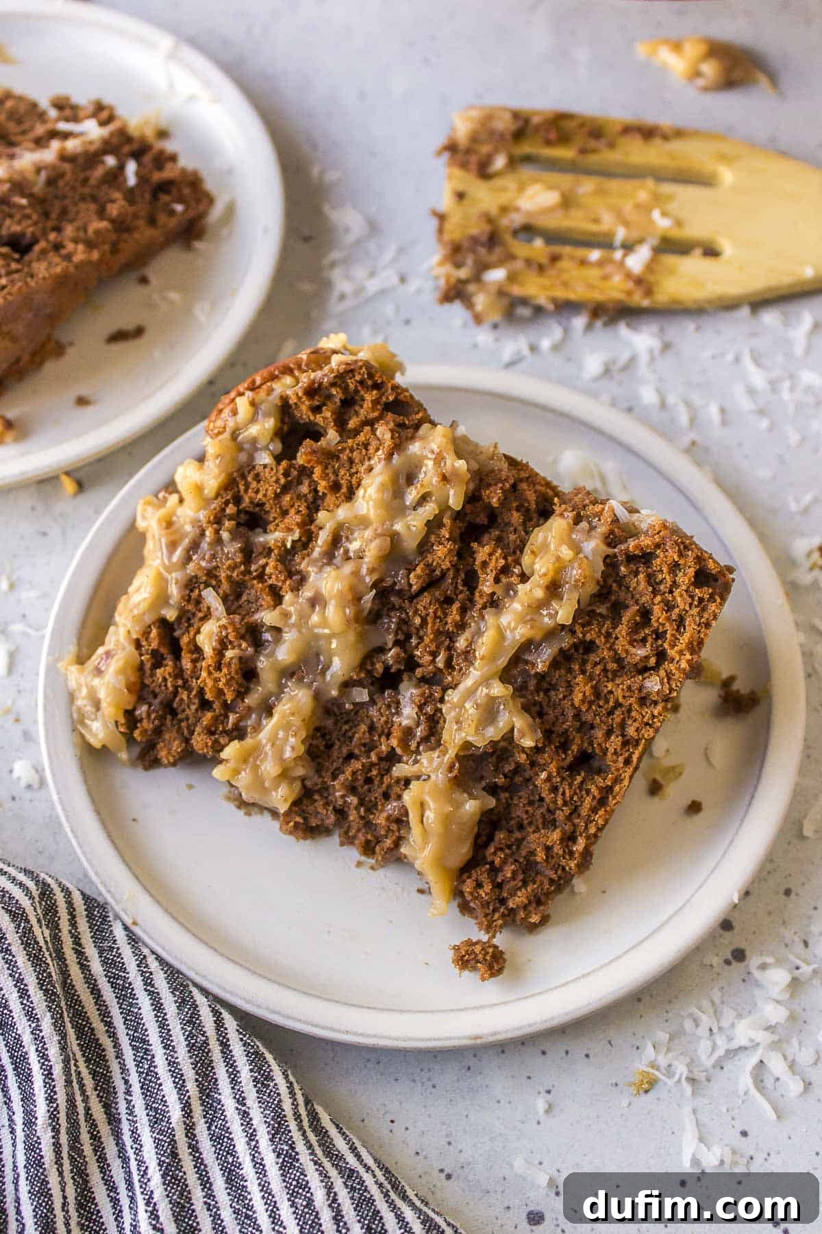 A slice of German chocolate cake resting on its side on a white plate, garnished with shredded coconut and surrounded by a stylish black and white striped kitchen towel. A wooden spatula and another cake slice in the background complete the inviting scene.