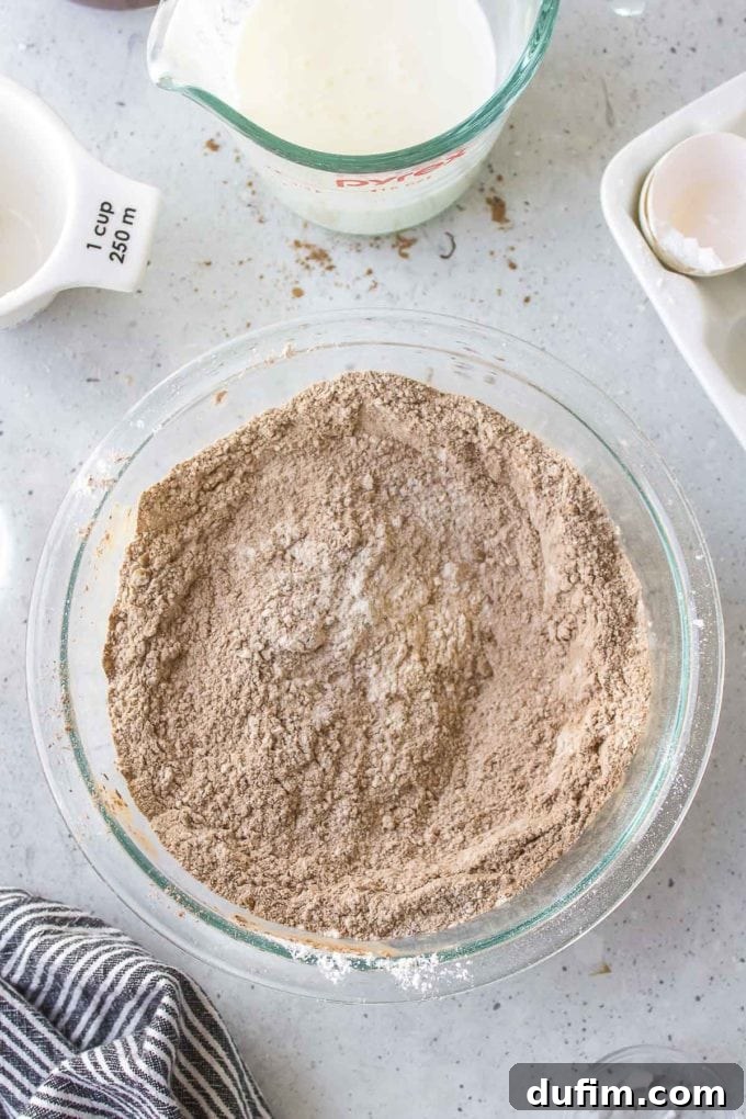 Ingredients for German chocolate cake in a glass bowl, next to a black and white striped towel, a white measuring cup, a glass measuring cup filled with buttermilk, and an empty eggshell in a white carton, showing the buttermilk ready to be added.