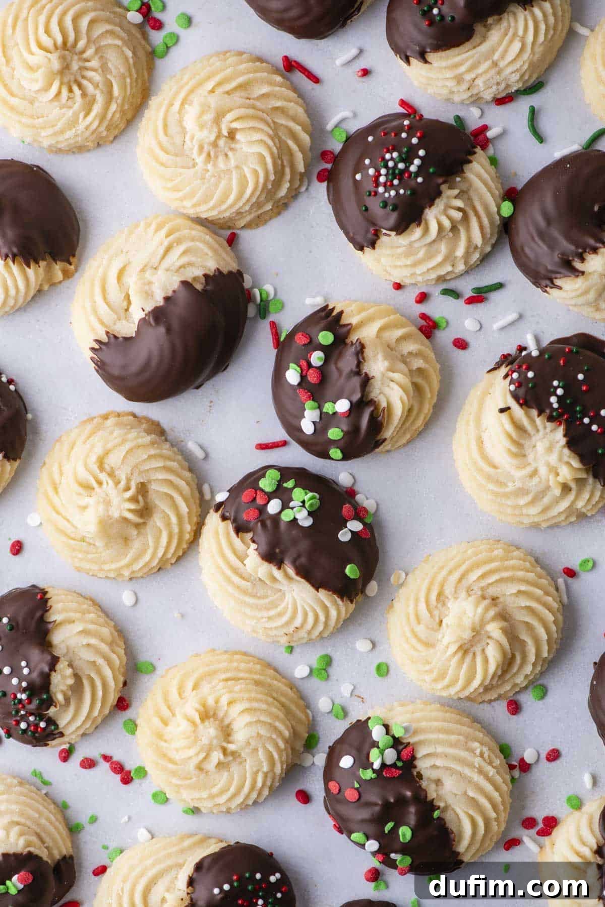 An assortment of golden butter cookies, some plain, some dipped in rich, dark chocolate, and others adorned with festive red, green, and white sprinkles, arranged neatly on parchment paper, with scattered sprinkles around.