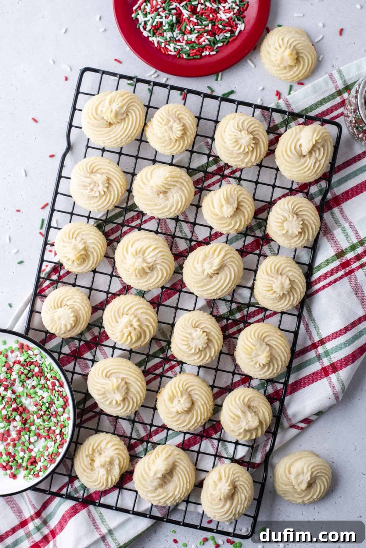 Plain butter cookies neatly arranged in rows on a wire rack, positioned over a festive red, white, and green kitchen towel. A small bowl of matching red, white, and green sprinkles is visible nearby, ready for decoration.