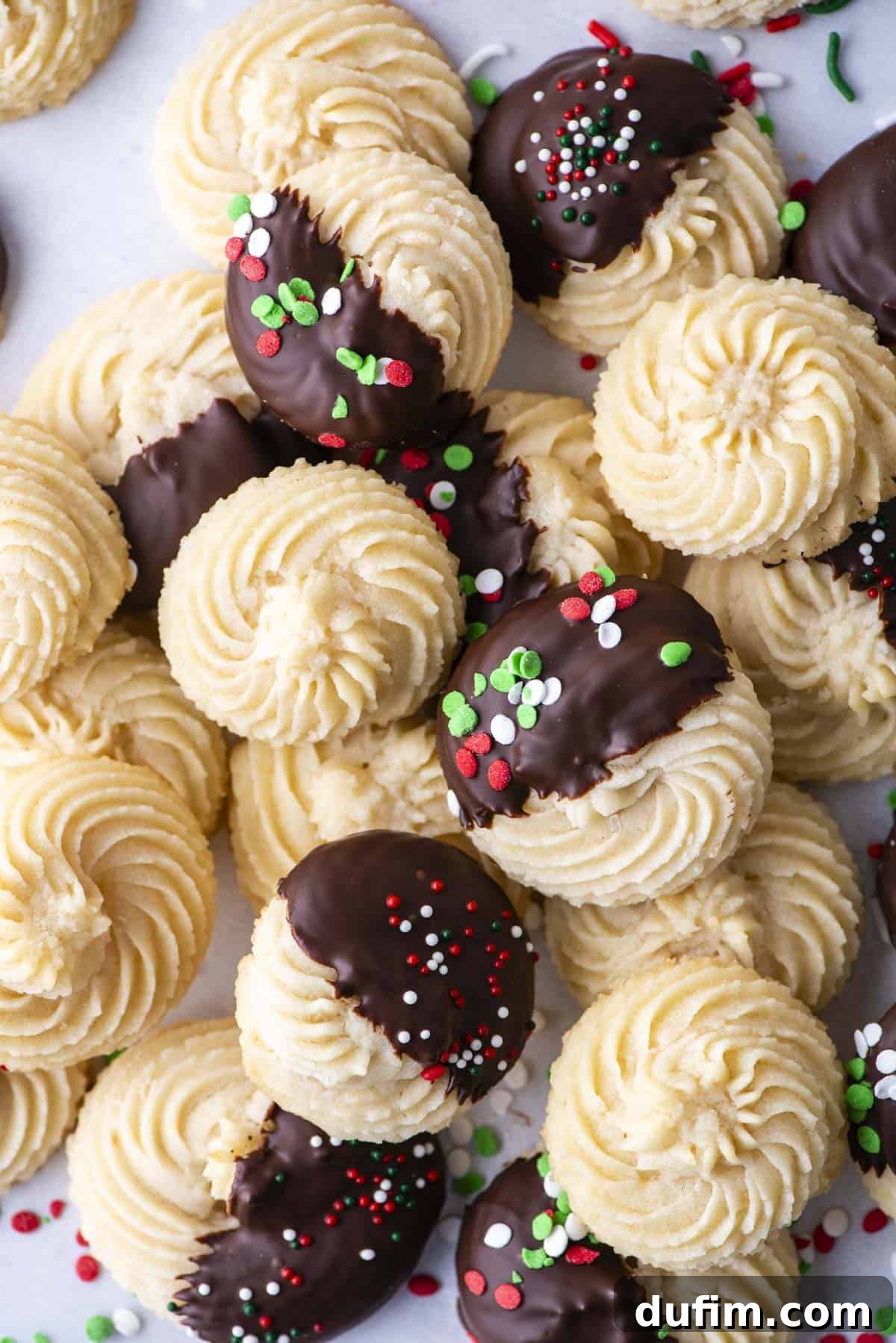 An inviting assortment of golden butter cookies, some plain, some expertly dipped in rich chocolate, and others charmingly decorated with festive red, green, and white sprinkles, beautifully piled on parchment paper.