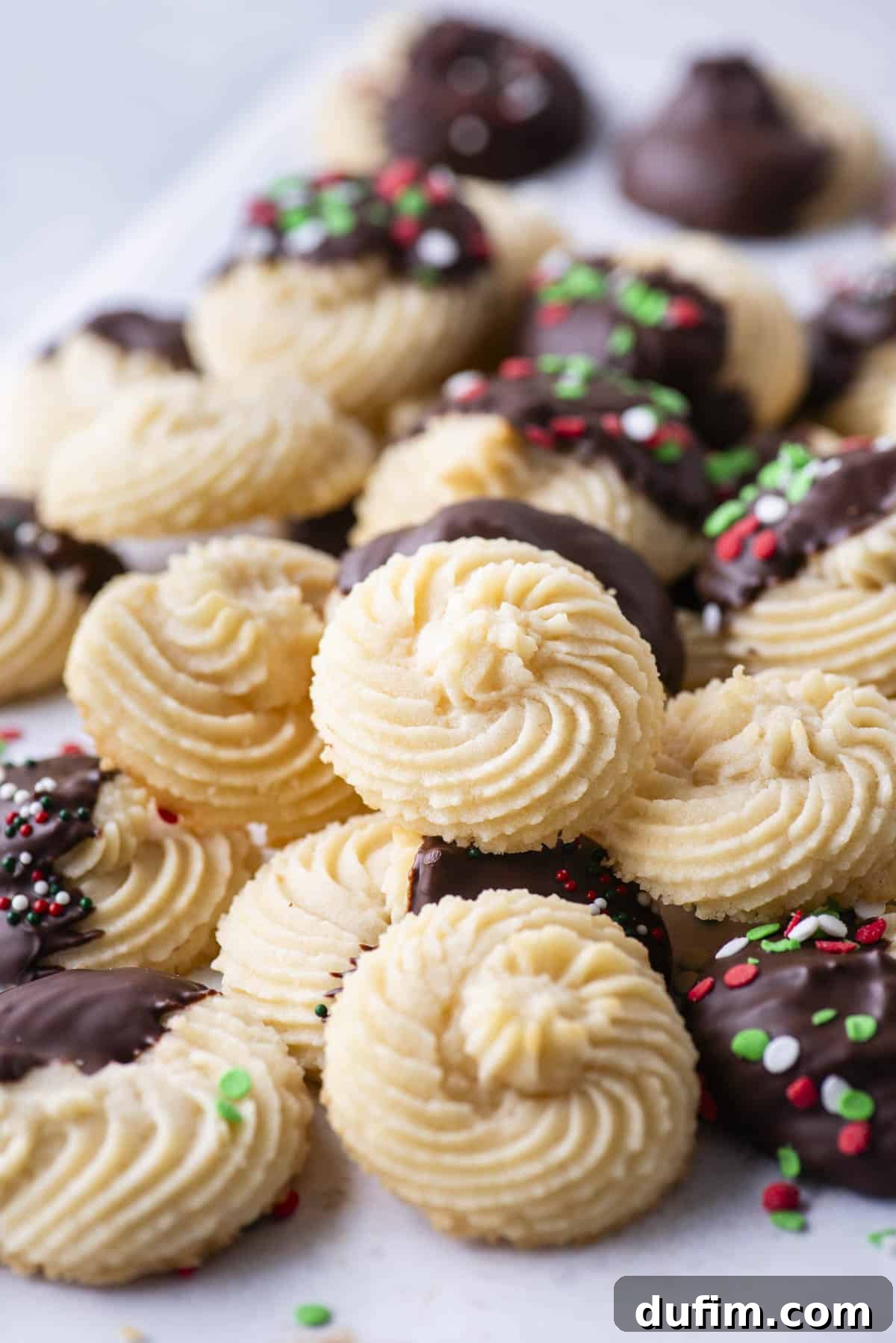 A delectable pile of butter cookies, featuring a mix of plain, chocolate-dipped, and sprinkle-decorated varieties, invitingly stacked on a surface.