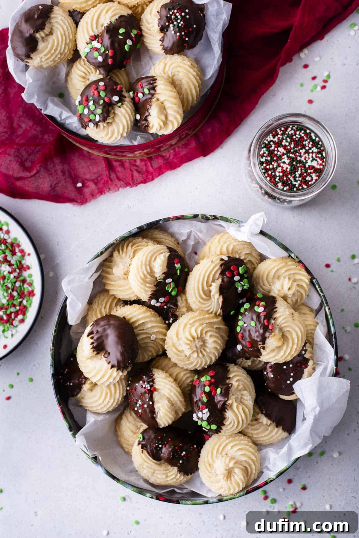 Two festive Christmas tins brimming with golden butter cookies, alongside a small bowl of colorful sprinkles. Some cookies are plain, others are dipped in chocolate, and many feature cheerful red, green, and white sprinkles, highlighting their holiday appeal.