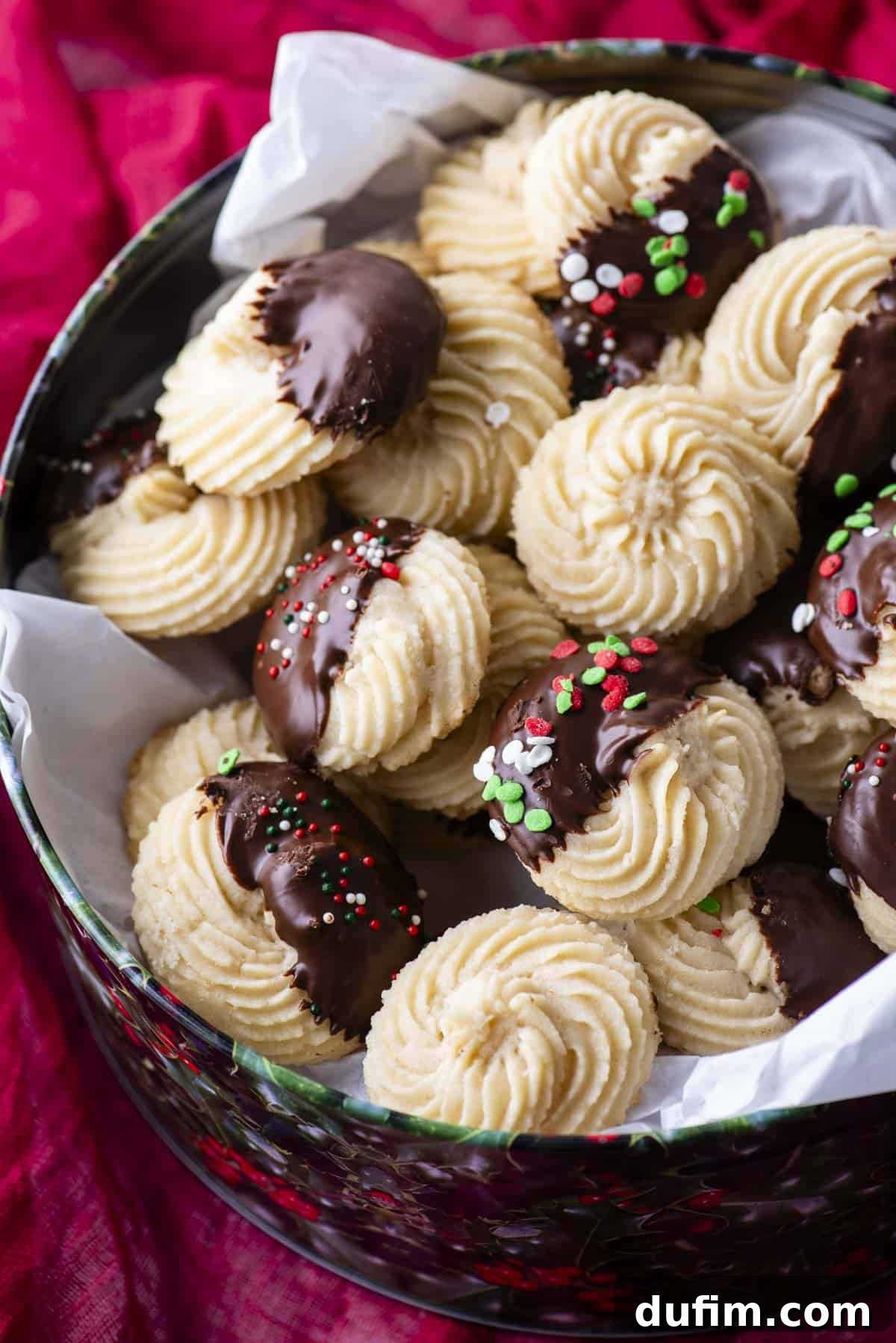 A festive Christmas tin filled to the brim with inviting butter cookies, featuring a mix of plain varieties and some half-dipped in rich chocolate, with a few adorned with cheerful red, green, and white sprinkles.