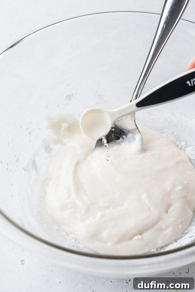 Royal icing in a clear glass bowl with a spoon and drops of water being added with a small measuring spoon.