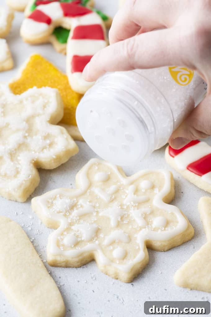 Snowflake cookies with white royal icing being dusted with sugar, with more Christmas-themed cookies in the background.