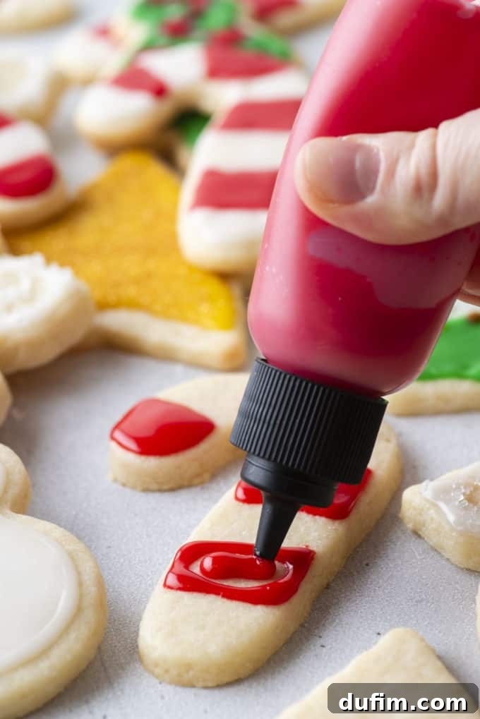 Red royal icing being piped onto a candy cane-shaped cookie using a squeeze bottle.