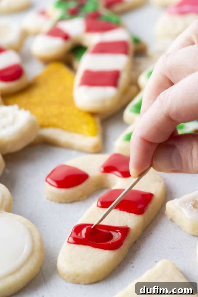 Red royal icing being spread using a toothpick on a candy cane-shaped cookie with more Christmas-themed cookies in the background.